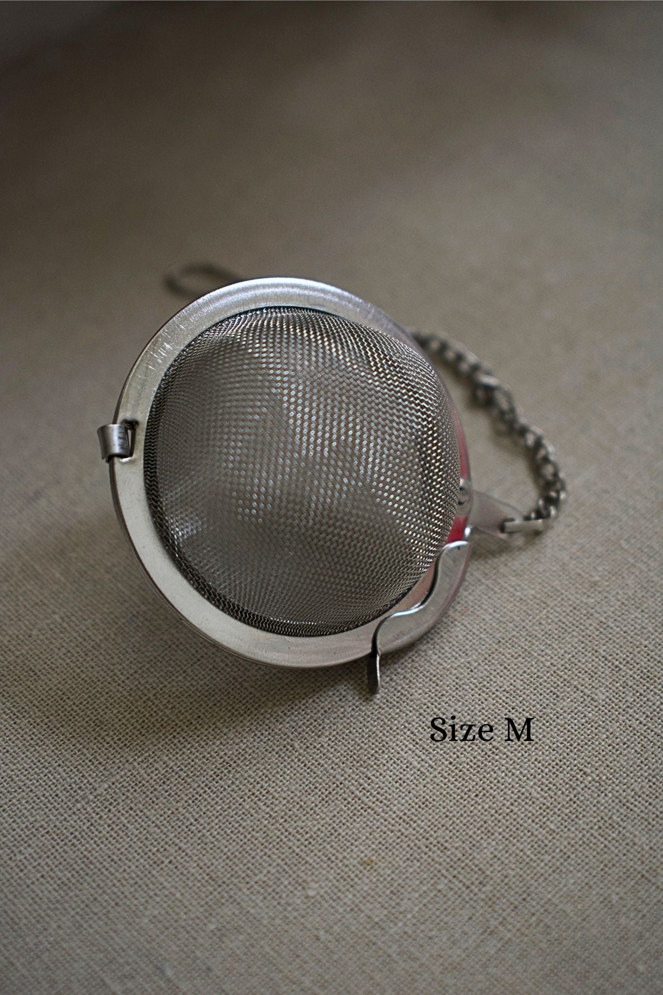 Metallic tea ball strainer on a textured surface with 'Size M' label.