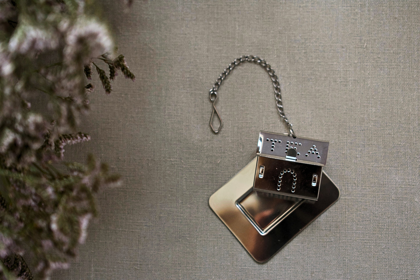 House shaped tea strainer with a chain, lying on little stainless steel plate. The whole is lying on linen tablecloth.
