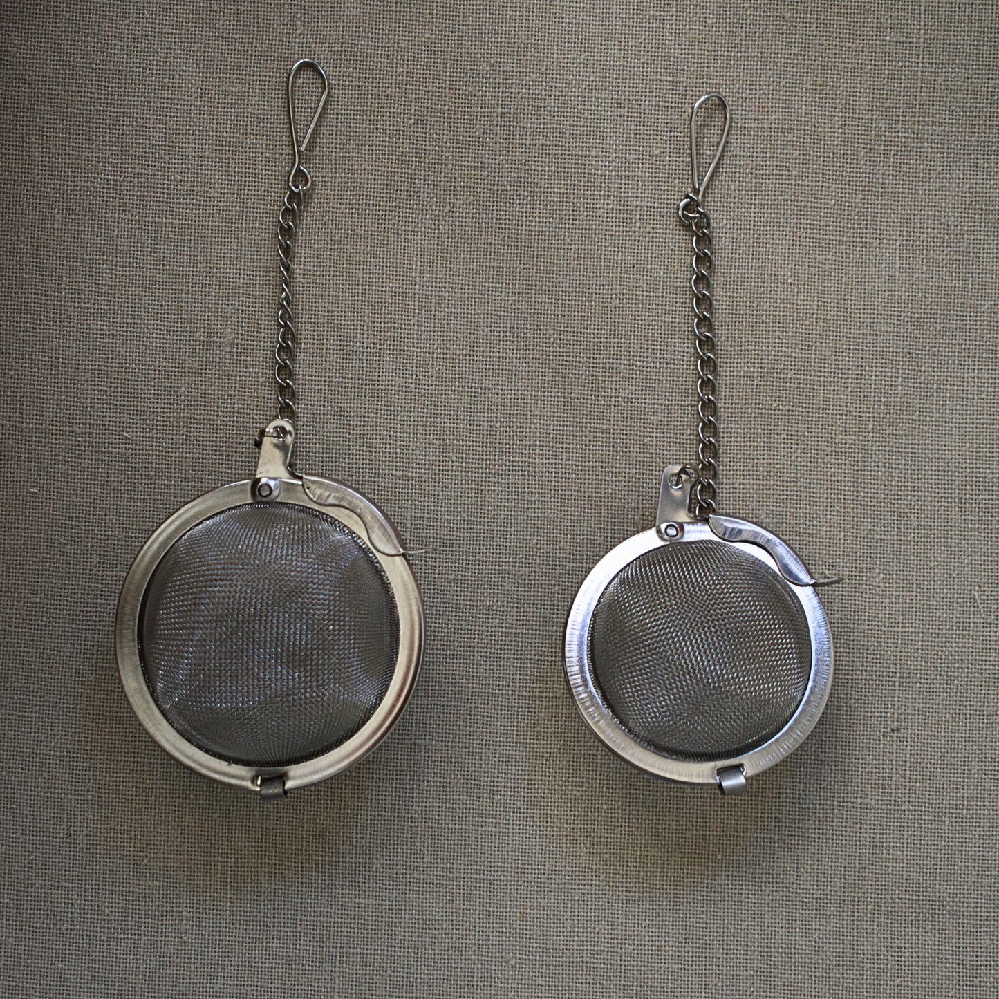 Two metal tea infusers with chains on a linen tablecloth. Size M and Size S