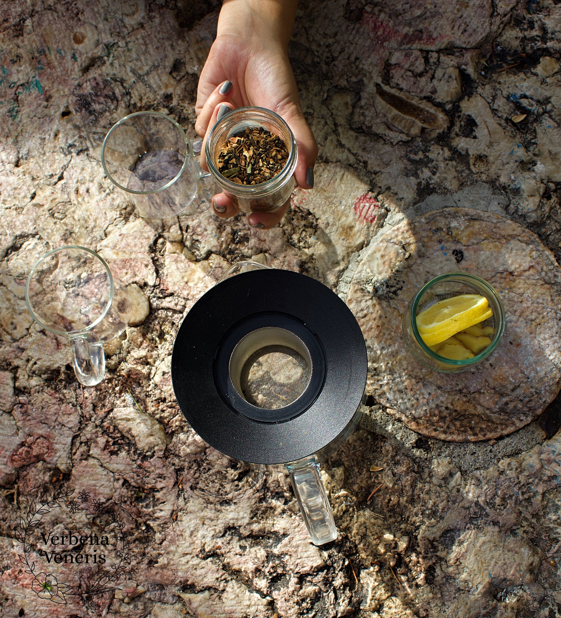 Context picture - tea pot and cup of  teas set  up on a table. A hand is holding a glass pot with the herbal blend Exotic Feeling