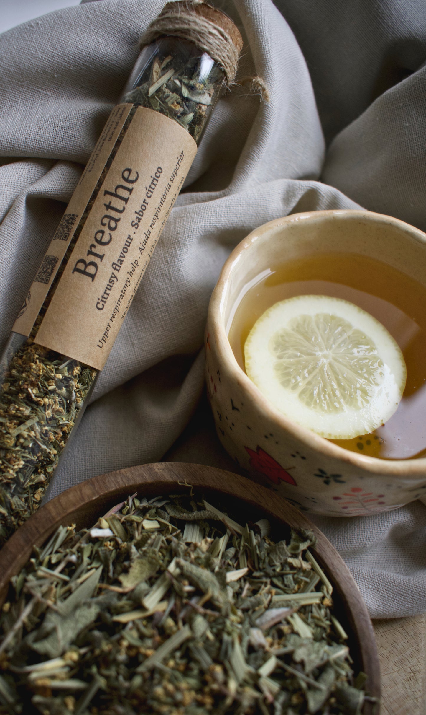 A glass tube with the tea blend Breathe, besides a cup of tea with a lemon slice and a wooden bowl with the tea blend Breathe. All elements are sitting on a linen tablecloth and a wooden board.