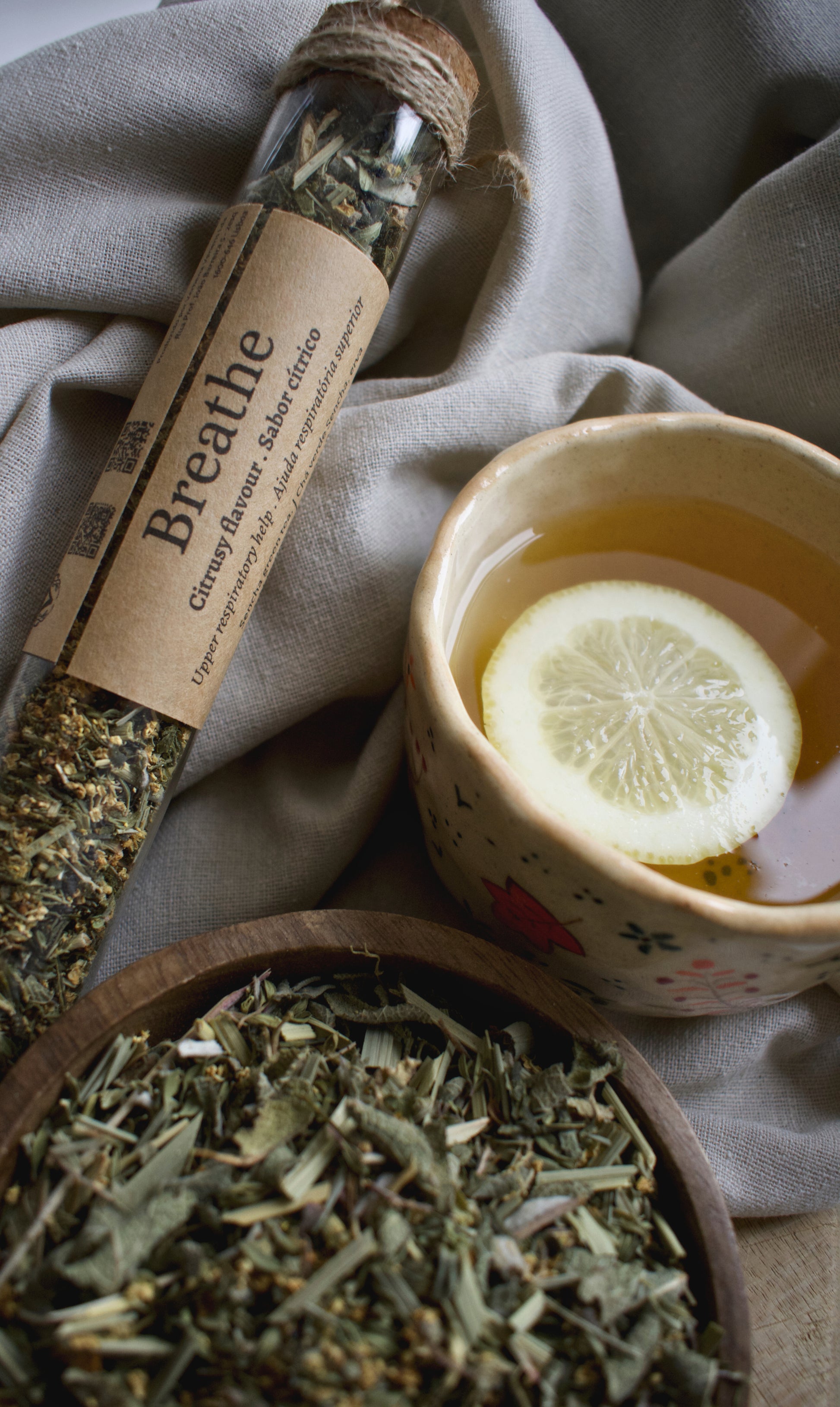 A glass tube with the tea blend Breathe, besides a cup of tea with a lemon slice and a wooden bowl with the tea blend Breathe. All elements are sitting on a linen tablecloth and a wooden board.