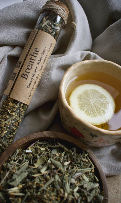 A glass tube with the tea blend Breathe, besides a cup of tea with a lemon slice and a wooden bowl with the tea blend Breathe. All elements are sitting on a linen tablecloth and a wooden board.