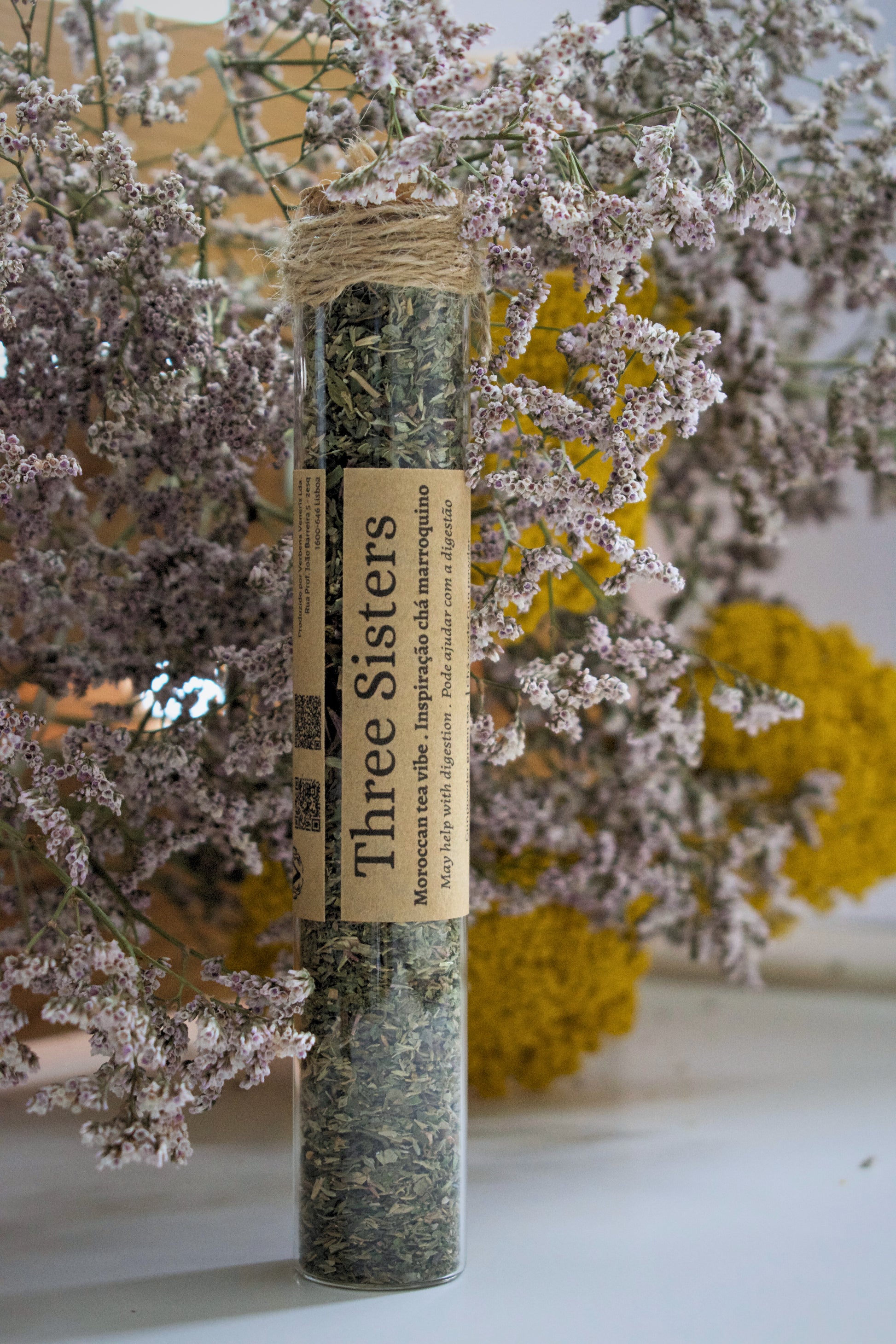 A glass tube with the tea blend Three Sisters standing up on a white surface, against a floral background