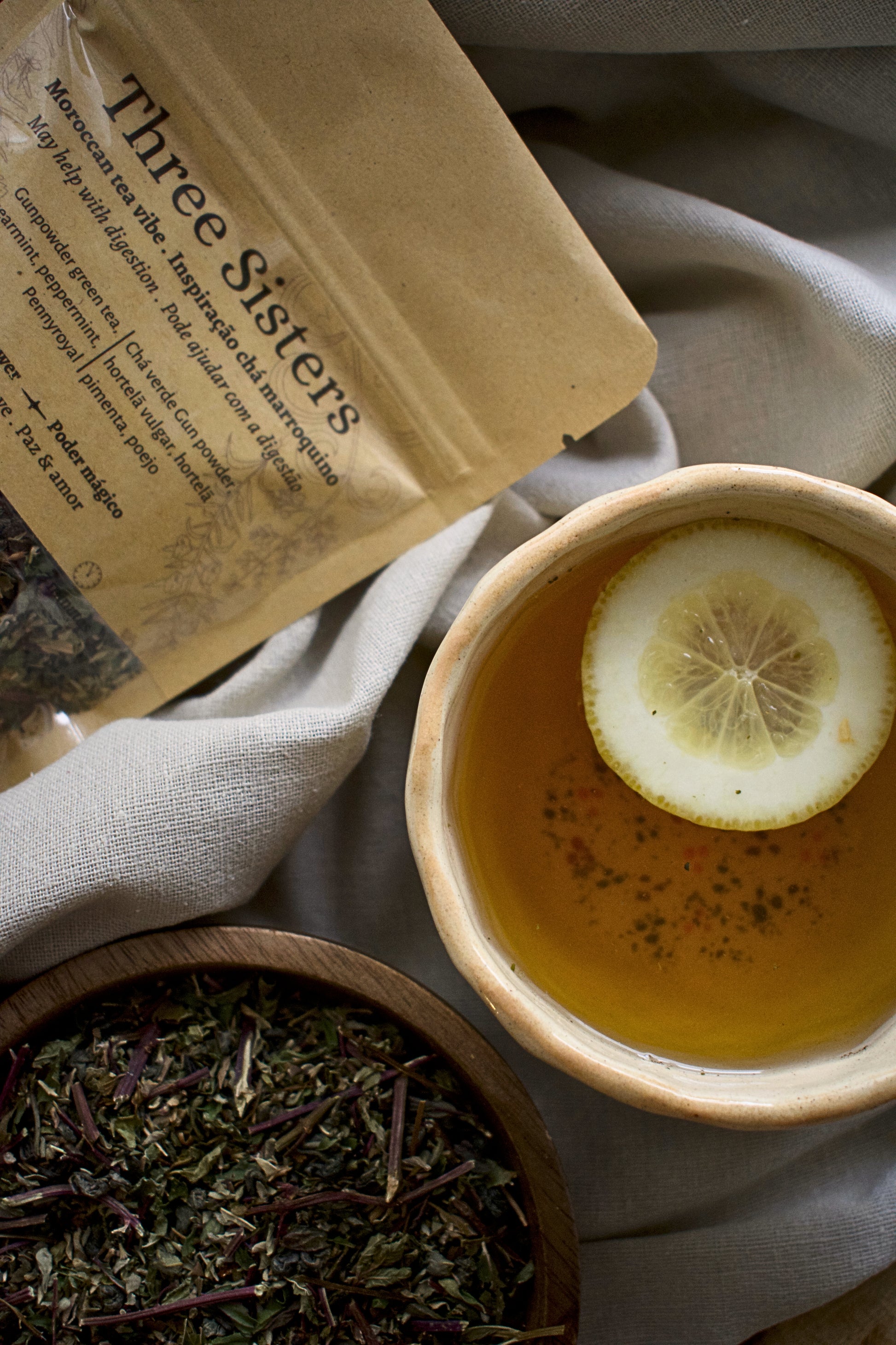A kraft bag of Three Sisters, , besides a cup of tea with a lemon slice and a wooden bowl with the tea blend Three Sisters. All elements are sitting on a linen tablecloth.