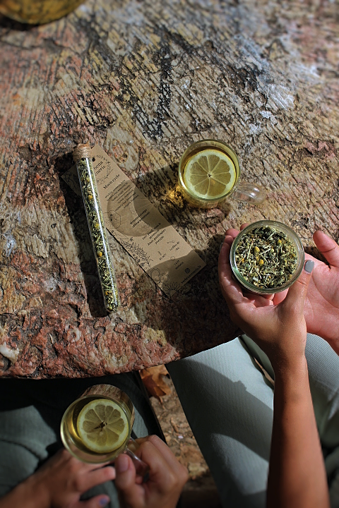 Context picture of moon cycle herbal blend. Set on a rock table we can see the chemist tube, two persons; one holding a cup of tea and the other a glass pot containing  moon cycle herbal blend