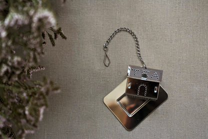 House shaped tea strainer with a chain, lying on little stainless steel plate. The whole is lying on linen tablecloth. 
