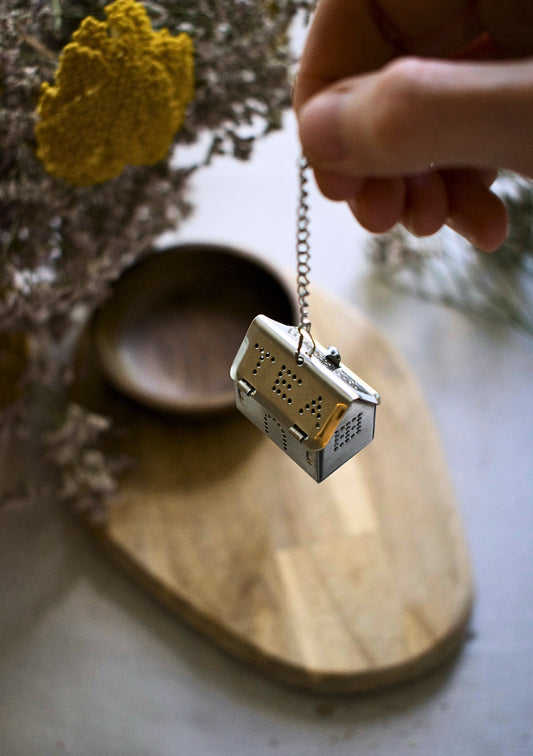 Hand holding a small house-shaped tea bag holder with a chain on a wooden surface.