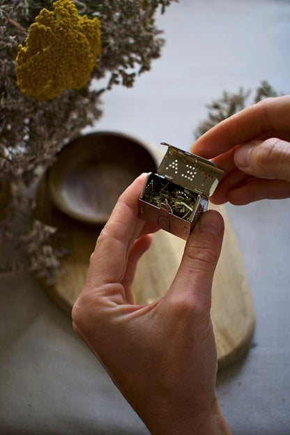Person holding a tea house shaped strainer with some tea inside, against a natural background.