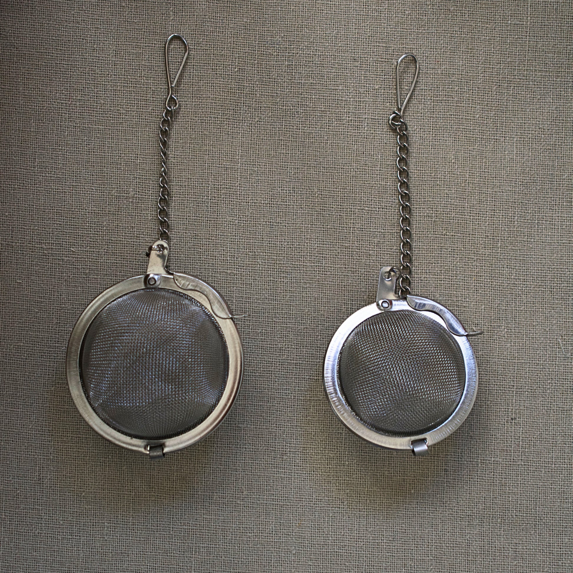Two metal tea infusers with chains on a linen tablecloth. Size M and Size S