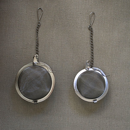 Two metal tea infusers with chains on a linen tablecloth. Size M and Size S